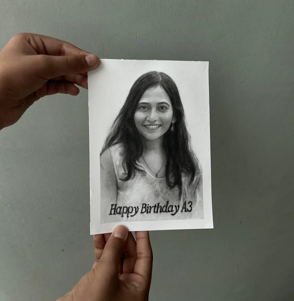A hand holds a printed black-and-white portrait card against a plain gray wall. The card shows a woman's face with the text 'Happy Birthday A3' at the bottom.