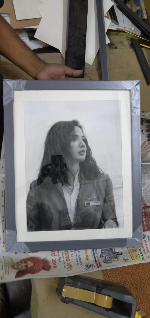 A framed black and white portrait print of a woman, held by a hand in a workshop or studio setting with papers and tools visible around it.