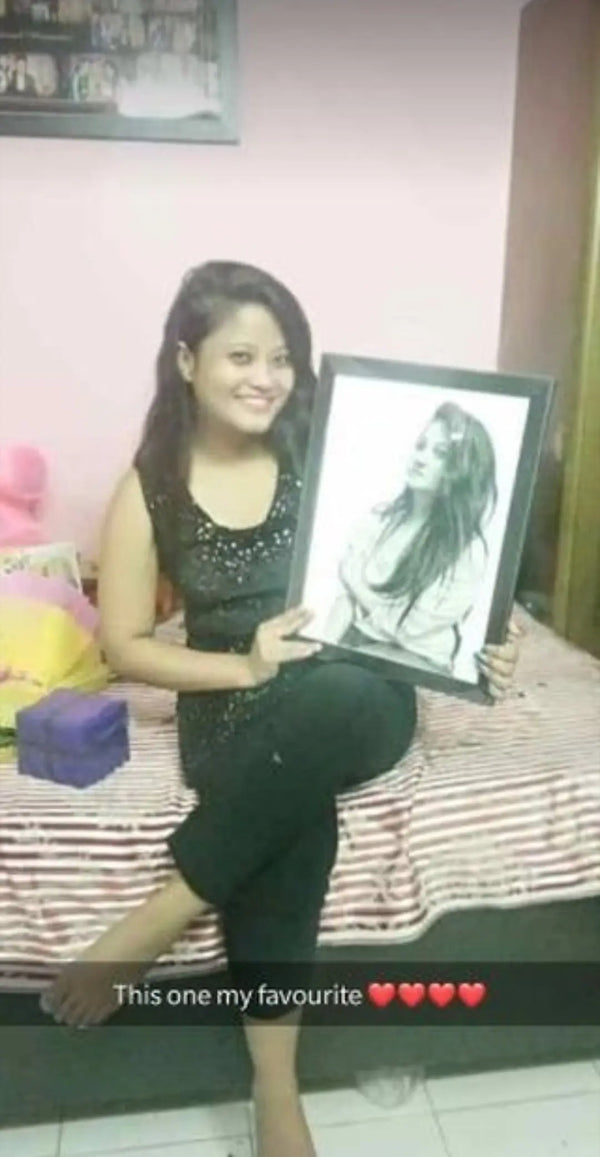 A woman sitting on a bed in a bedroom holds up a framed black-and-white portrait sketch of a woman. The room has pink walls, a striped bedspread, and plush toys on the left side.