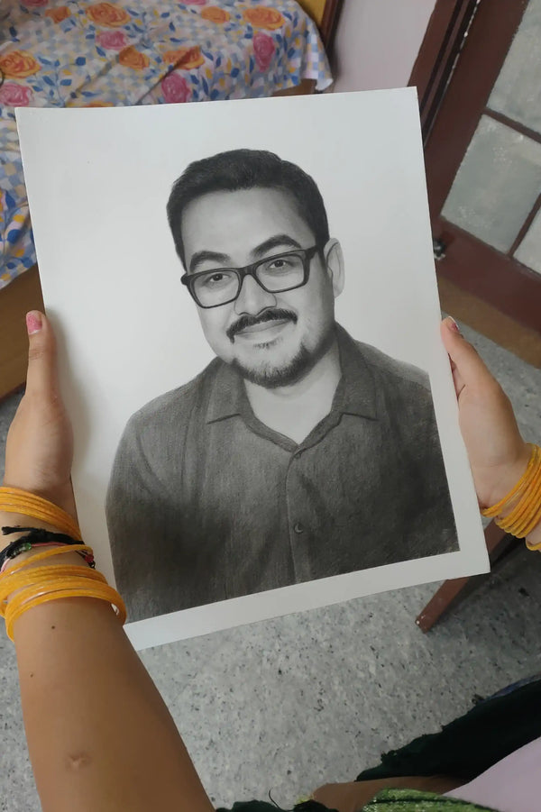 A person holds up a black and white pencil sketch portrait of a man with glasses and a mustache. The person's hands, adorned with yellow bangles, are visible holding the drawing paper in an indoor setting.