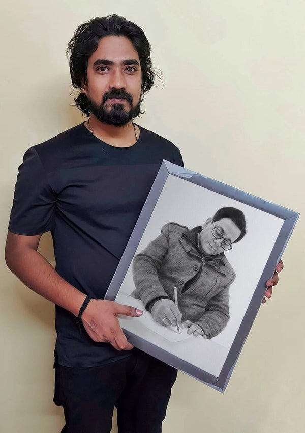 A man with a beard and dark long hair wearing a black t-shirt holds up a framed black and white pencil sketch portrait of a man writing. The background is a plain beige wall.