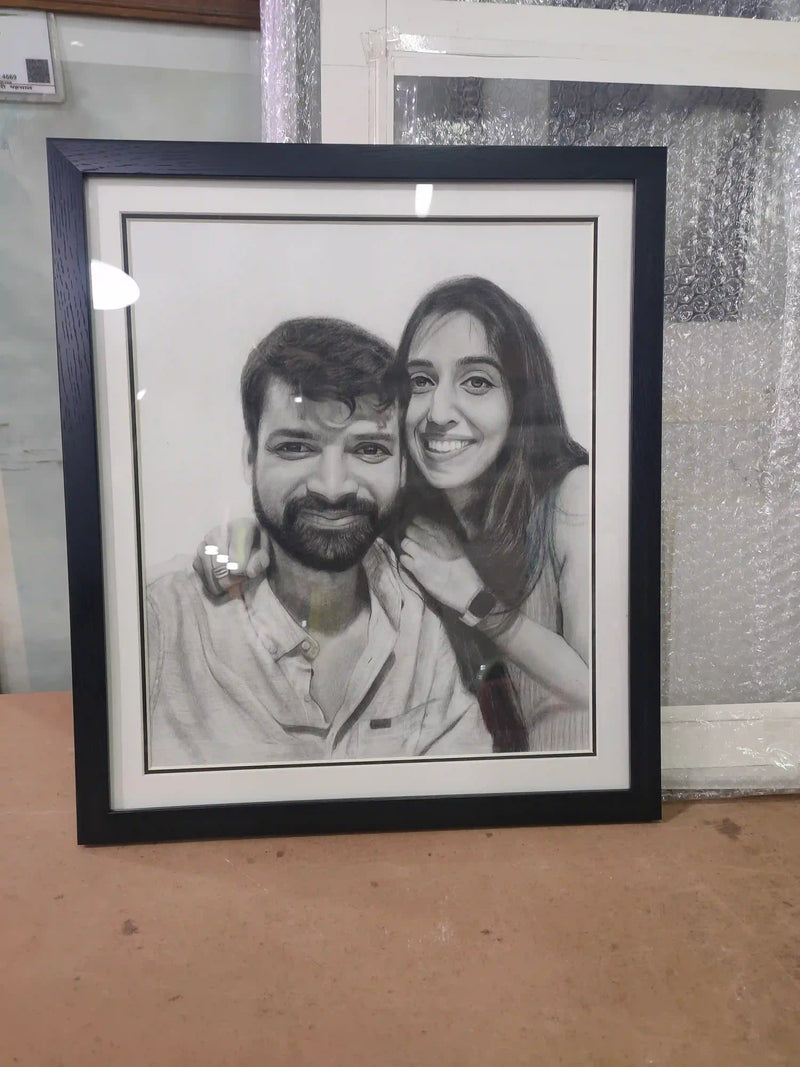 A black-framed custom pencil sketch portrait of a smiling couple, displayed on a wooden surface with a bubble-wrapped glass window in the background.