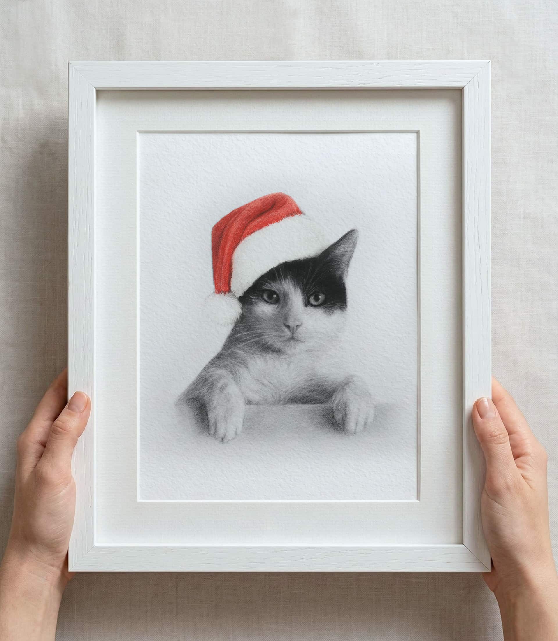 Custom black and white pencil sketch portrait of a cat wearing red colour Santa hat in a white frame with white mount board, held by hands on a light background. 
