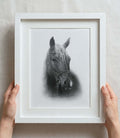 Black and white custom horse drawing in a white frame with white mount board, held by two hands on a light background.